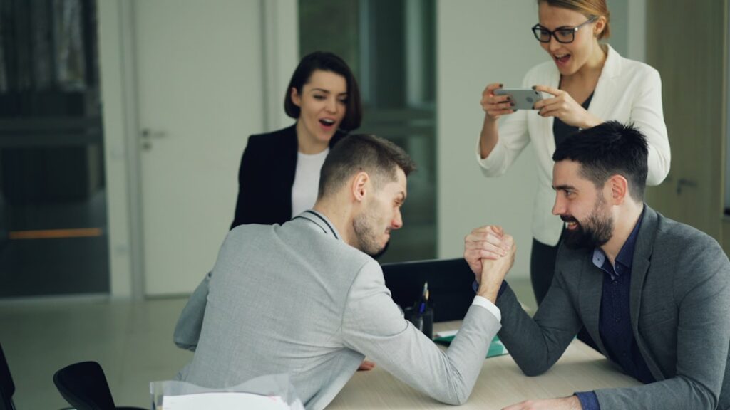 Colleagues arm wrestling while others watch and film.